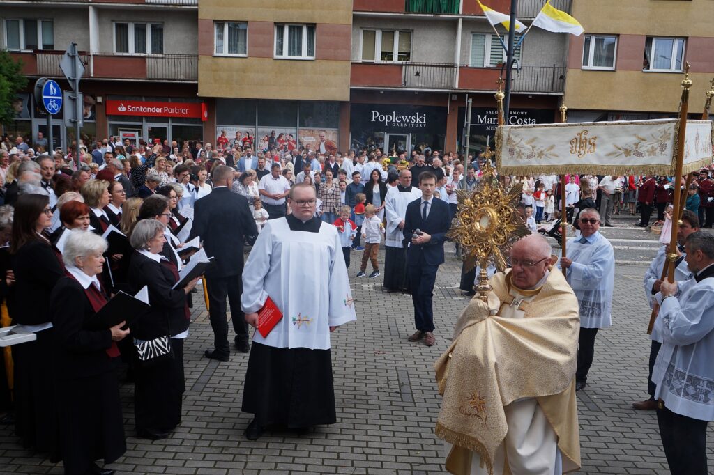 Tłum wiernych w procesji. Kapłan celebrans niesie Monstrancję z Hostią. 
