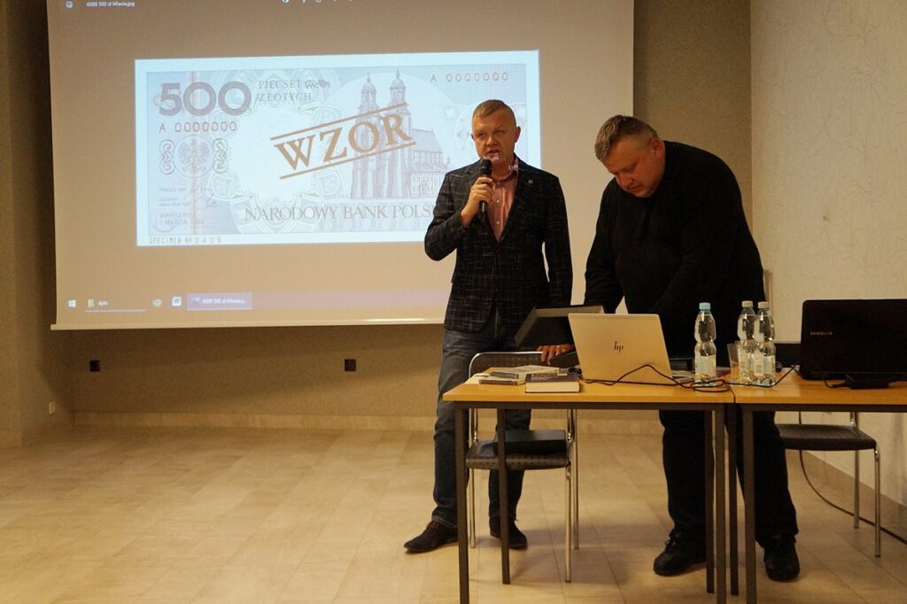 Dwóch mężczyzn: Prezes  Polskiego Towarzystwa Numizmatycznego Oddział w Jaśle i ksiądz Marek Dyło, prezentują kolekcję banknotów i medali.