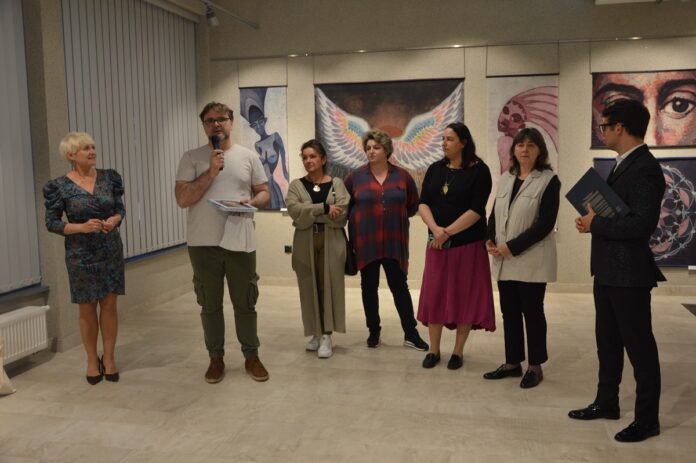 Siedem osób: pięć kobiet i dwóch mężczyzn. Jeden z mężczyzn trzyma mikrofon w ręku i przemawia. Wszyscy uczestniczą w uroczystej inauguracji wystawy.