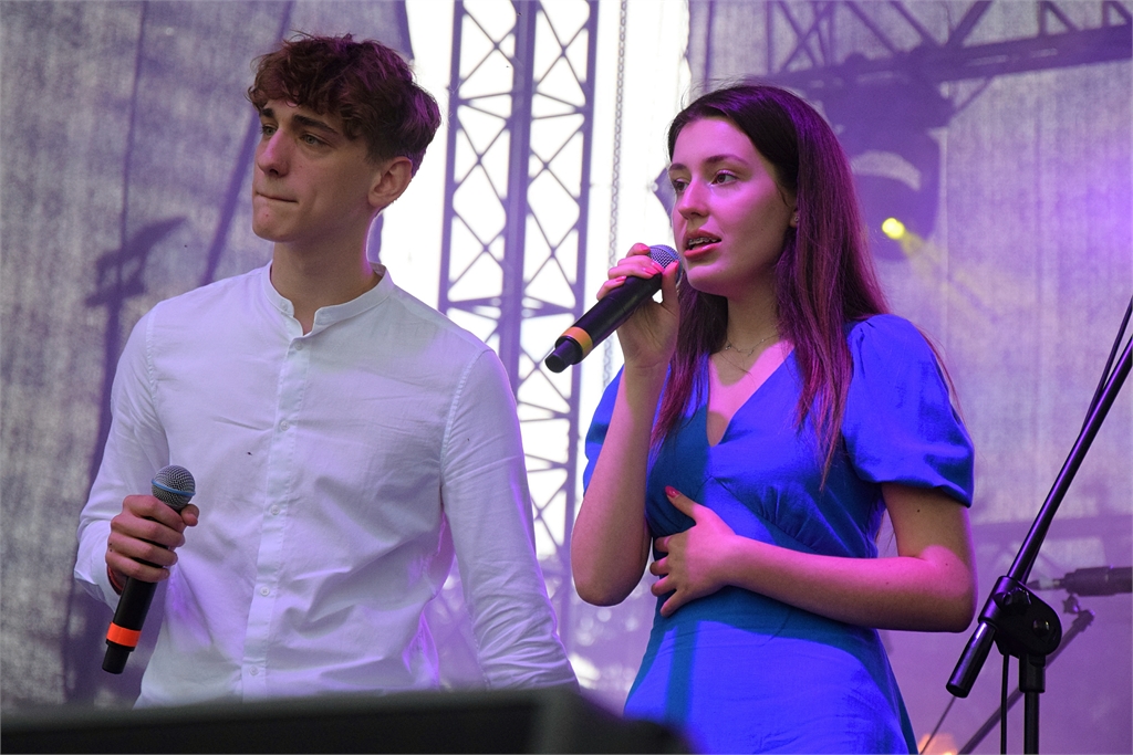 Obraz zawiera: Młodzi wykonawcy stoją na koncertowej scenie plenerowej. Młody mężczyzna z lewej strony trzyma w prawej dłoni mikrofon na wysokości klatki piersiowej i patrzy w dal. Młoda kobieta z prawej strony trzyma lewą dłoń na przeponie, a w prawej trzy mikrofon na wysokości ust i śpiewa.
