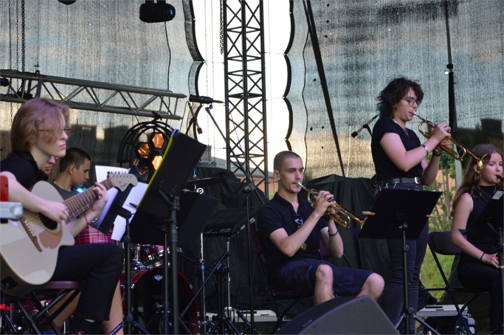 Obraz zawiera: Muzycy formacji Jazz of Combo na scenie. Z lewej strony dwaj gitarzyści, perkusista oraz sekcja dęta. Młoda trębaczka w okularach stoi, wykonując partię solową.