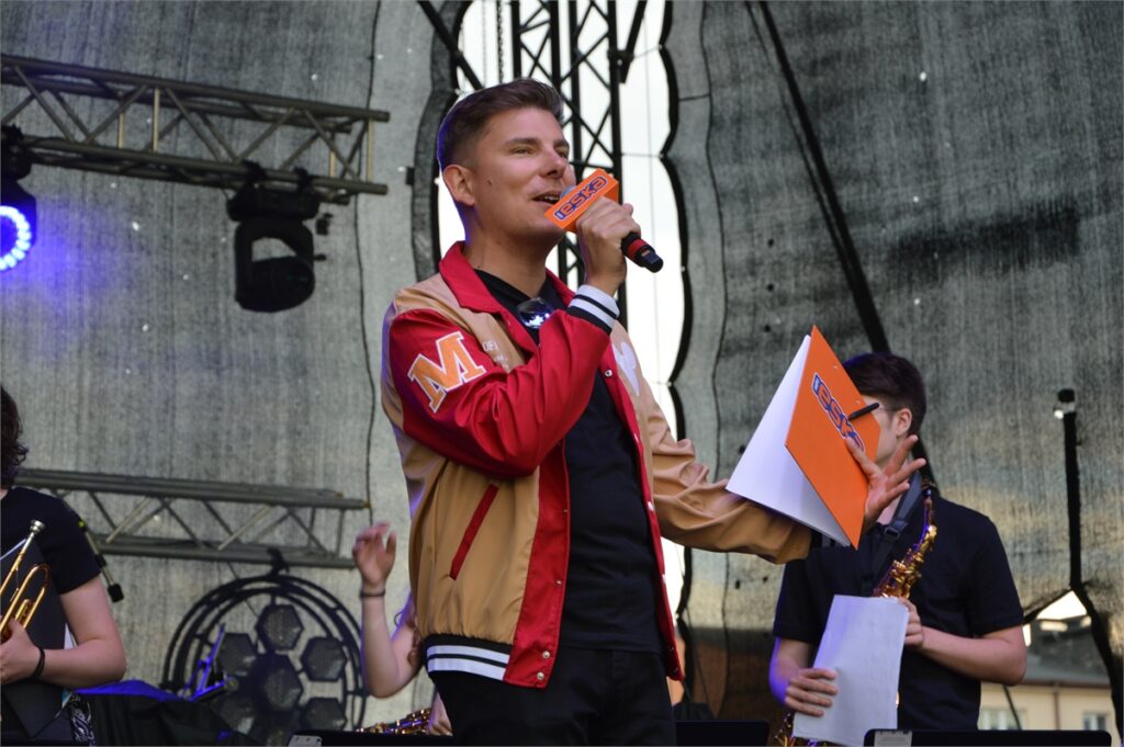 Obraz zawiera: Młody mężczyzna w sportowej bluzie męskiej rozpinanej stoi na scenie i mówi do trzymanego w rękach mikrofonu. Na mikrofonie nałożona jest kostka z logo Radia ESKA. w lewej ręce trzyma również skoroszyt z logo Radia ESKA.