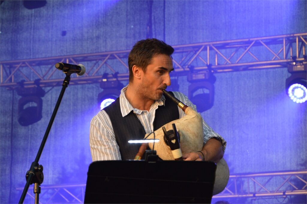 Obraz zawiera: Mężczyzna spogląda w prawo i gra na dudach. Przed nim podświetlona tablica z nutami. Z jego lewej strony stoi statyw z mikrofonem.