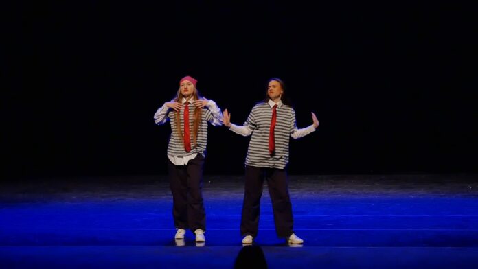 Duet 02 Dwie, młode dziewczyny tańczą na scenie. Ubrane są w szare koszule w czarne paski, długi czerwony krawat, czarne spodnie i białe buty.