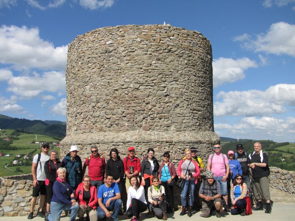 Grupa osób na szczycie wzgórza. Wszyscy pozują do zdjęcia. Stoją przed fragmentem wieży zamkowej. 