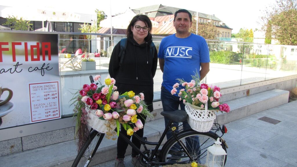 Dwie osoby: kobieta i mężczyzna, pozują do zdjęcia. Przed nimi czarny rower z dwoma białymi koszami, w których znajdują się kolorowe kwiaty. 