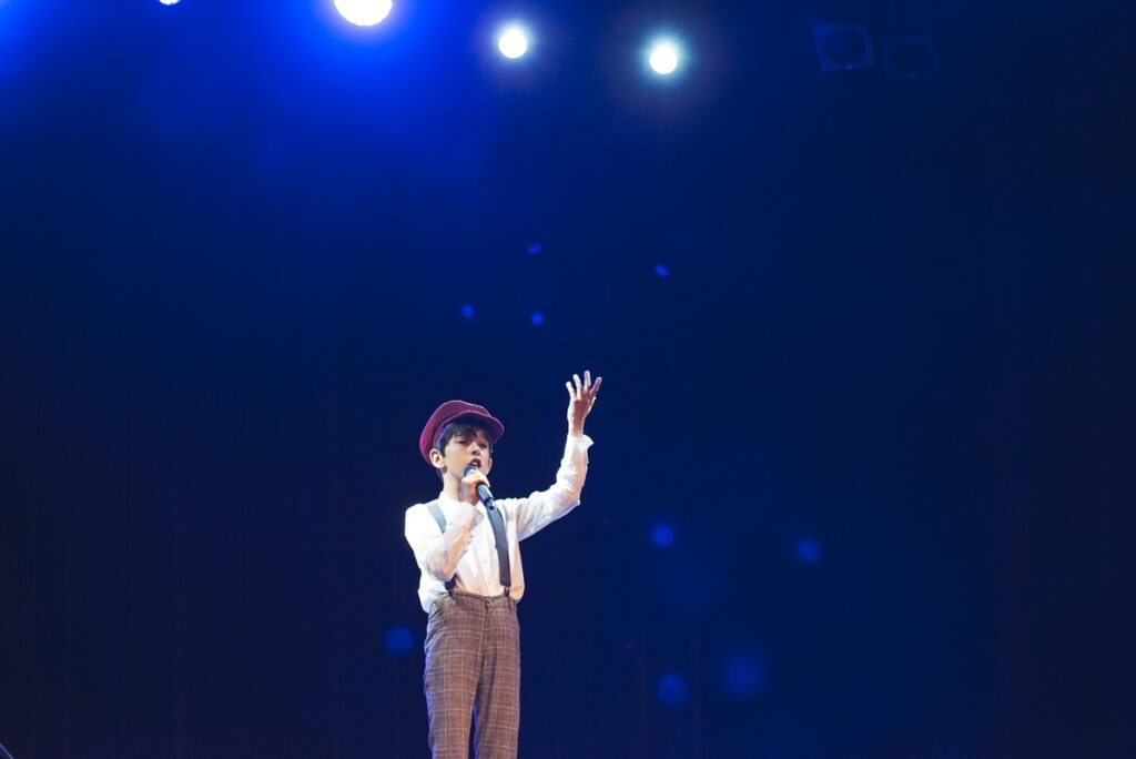 Młody chłopak śpiewa na scenie. W dłoni trzyma mikrofon. Ma na sobie białą koszulę, jasno brązowe spodnie w delikatną kratkę i czerwoną czapkę z daszkiem na głowie. 