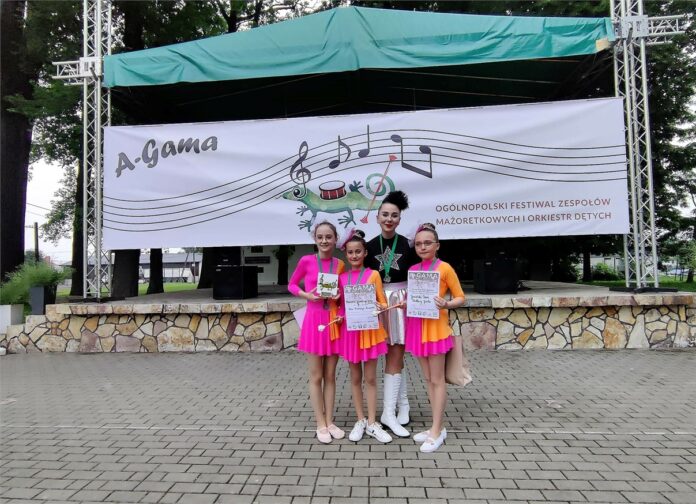 Obraz zawiera: Cztery osoby stoją przed sceną plenerową Ogólnopolskiego Festiwalu Zespołów Mażoretkowych i Orkiestr Dętych A-Gama. Na scenie rozwieszony jest baner tego wydarzenia. Trzy dziewczynki w strojach mażoretek pozują do zdjęcia z dyplomami i statuetką przeglądu, którą trzyma dziewczynka pierwsza z lewej strony. Tuż za dziewczynkami pozuje do zdjęcia ich opiekunka. Wszystkie mają upięte włosy oraz medale na szyi.