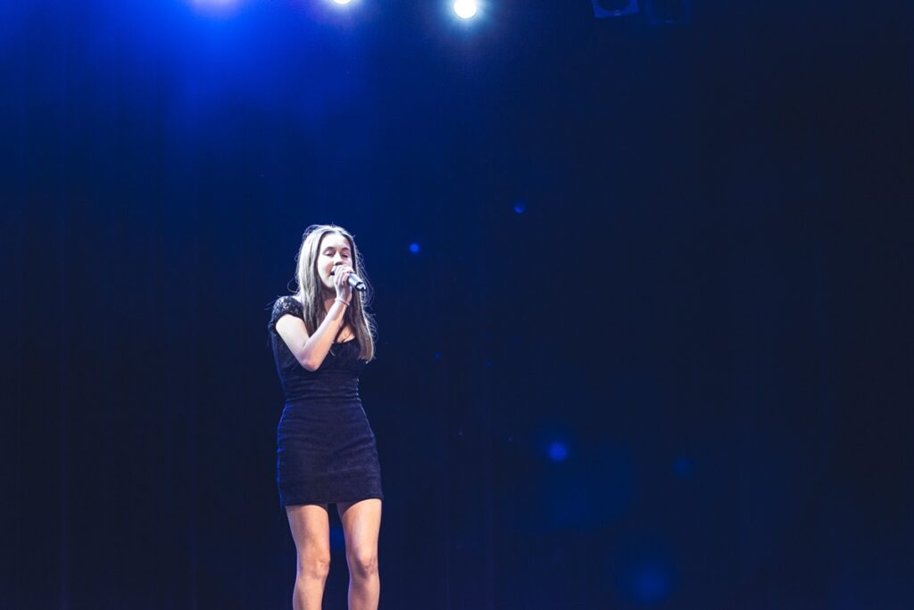 Młoda dziewczyna śpiewa na scenie. W dłoniach trzyma mikrofon. Ubrana jest w czarną sukienkę. 