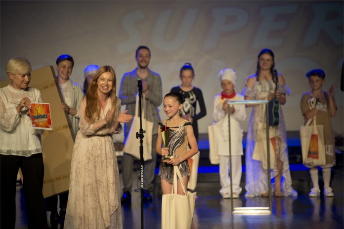 Wręczenie Super Talenciaka_11 Obraz zawiera: Na pierwszym planie dziewczynka w stroju akrobatki ze statuetką Supertalenciaka w rękach i torbą z upominkami. Obok niej brawo bije Elwira Musiałowicz - Czech, zastępca burmistrza Jasła, z jej lewej strony stoi dyrektor placówki - Helena Gołębiowska. Z tyłu za nimi stoją Katarzyna Korona - pomysłodawca konkursu trzymająca w ręku nagrodę dla laureatki, a także pozostali finaliści konkursu, którzy także biją brawo zwyciężczyni.