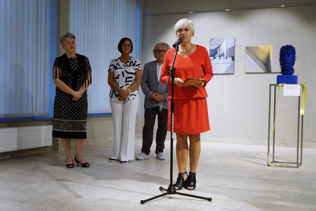 Cztery osoby: trzy kobiety i jeden mężczyzna. Kobieta w czerwonej sukience, czyli Pani Dyrektor Jasielskiego Domu Kultury Helena Gołębiowska, przemawia do mikrofonu, witając serdecznie przybyłych gości.