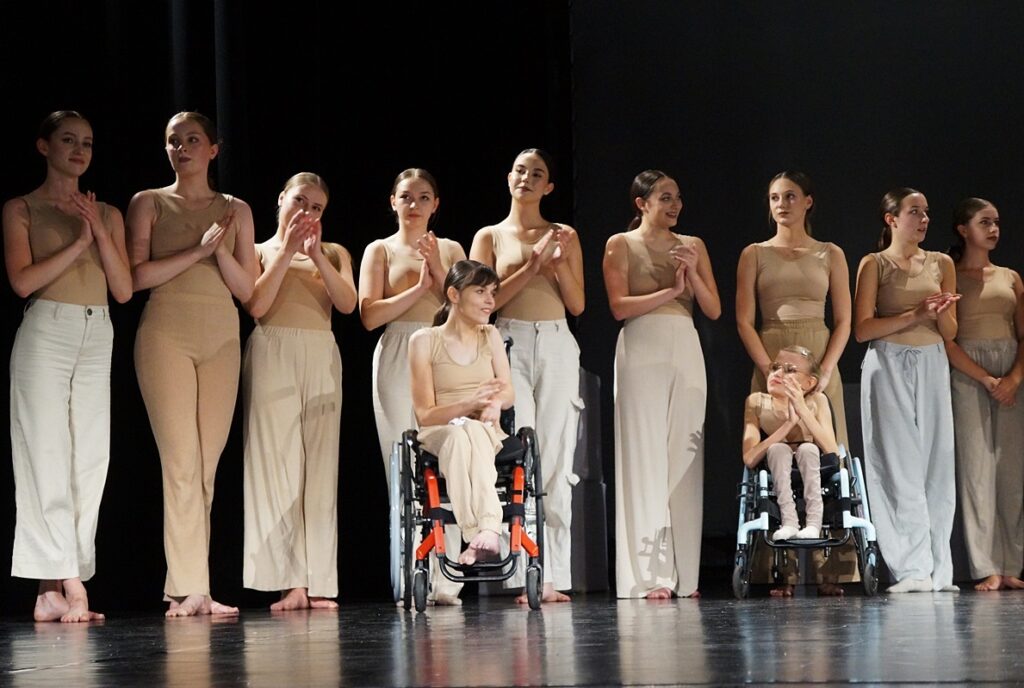 Grupa dziewcząt stoi na scenie. Dwie tancerki na wózku inwalidzkim. 