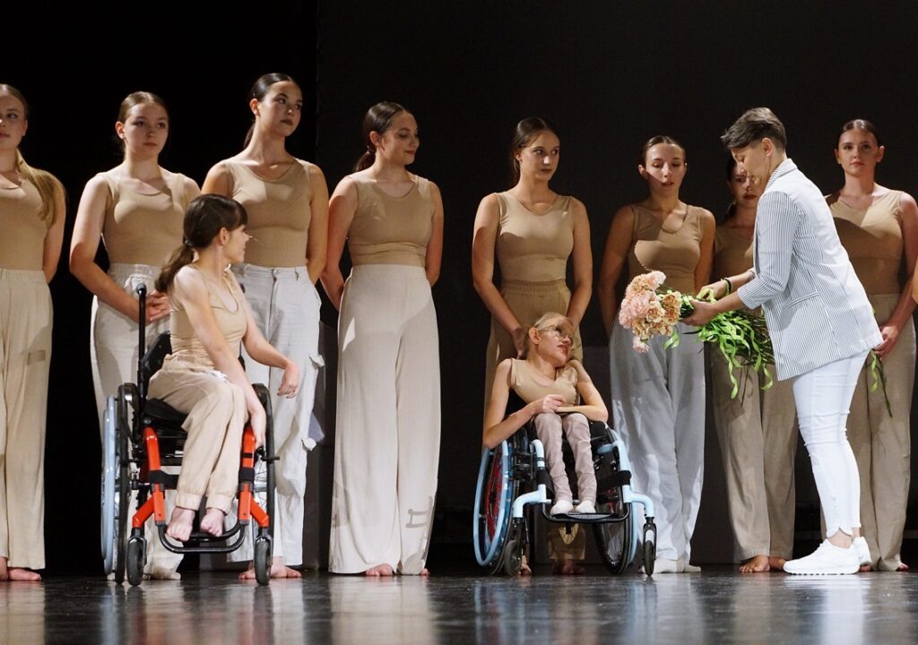 Grupa dziewcząt na scenie. Każda z tancerek otrzymuje kwiat (różową różę) z rąk Pani Katarzyny Korony, instruktor tańca w Jasielskim Domu Kultury. Dwie tancerki na wózku inwalidzkim. 
