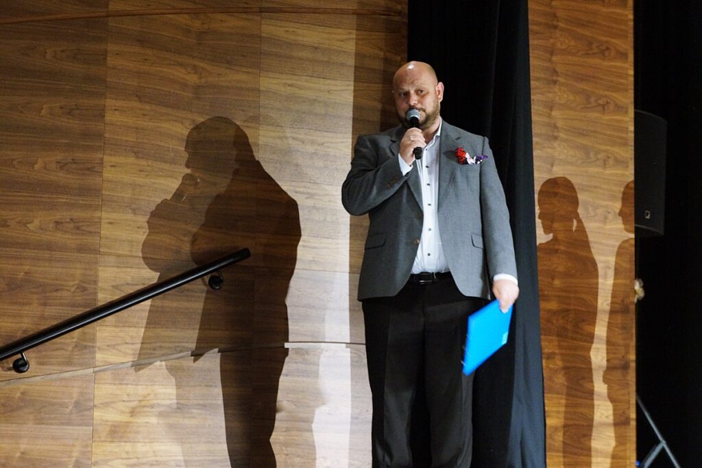 Na scenie stoi mężczyzna, który przemawia do mikrofonu. Ubrany jest w białą koszulę, szarą marynarkę i czarne spodnie. W lewej dłoni trzyma niebieską teczkę. 
