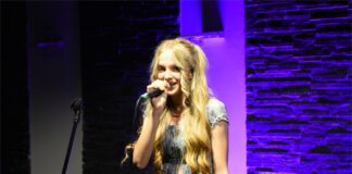 Koncert z Bossa novą w tle Obraz zawiera: Młoda dziewczyna z jasnymi lekko falującymi włosami opadającymi do pasa śpiewa do mikrofonu trzymanego w prawej ręce i uśmiecha się lekko. Ma na sobie jasną sukienkę z ramionami.