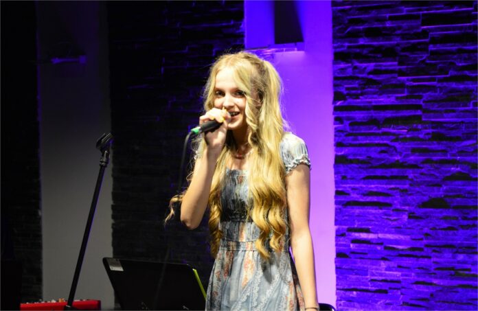 MB Bossa nova_03 Obraz zawiera: Młoda dziewczyna z jasnymi lekko falującymi włosami opadającymi do pasa śpiewa do mikrofonu trzymanego w prawej ręce i uśmiecha się lekko. Ma na sobie jasną sukienkę z ramionami.