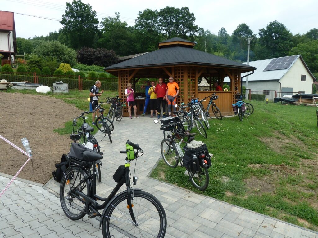 Grupa rowerzystów, stoi przed drewnianą altanką. 