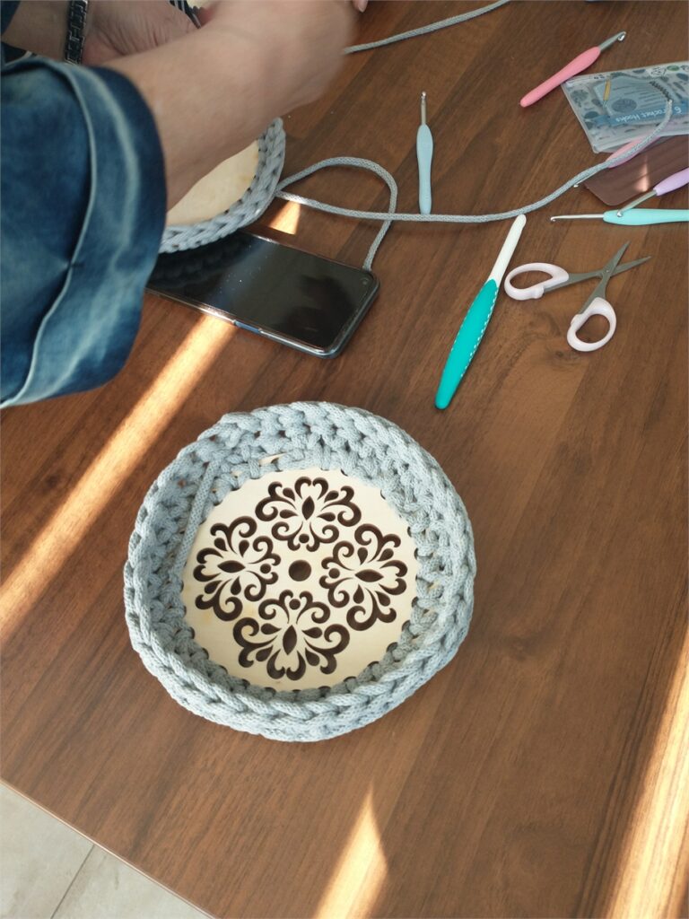 Obraza zawiera: Na stole koszyczek w trakcie tworzenia obok szydełka, nożyczki i smartfon twórcy.