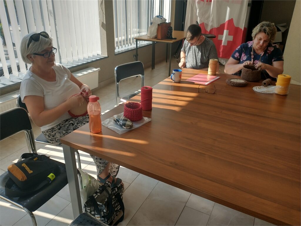 Obraz zawiera: Trzy osoby, dwie panie i jeden pan szydełkują przy stole. Naprzeciwko każdej z osób stoją szpulki nici potrzebne do wykonywania prac.