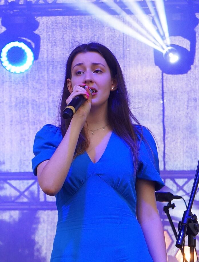 Polak Młoda artystka: Karolina Polak, śpiewa do mikrofonu. Ubrana jest w błękitną, letnią, zwiewną sukienkę.