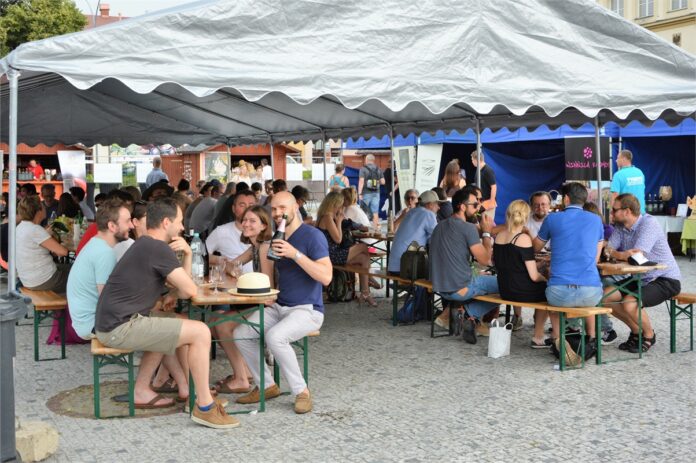 Obraz zawiera: Namiot degustacyjny produktów zakupionych podczas XVII Międzynarodowych Dni Wina. Pod nim na ławkach, przy stolikach siedzą osoby przybyłe na wydarzenie, degustujące zakupione produkty.