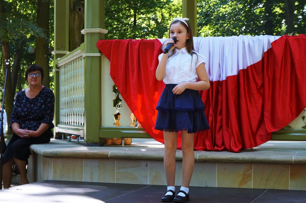 Mała dziewczynka stoi na scenie. W dłoni trzyma mikrofon i śpiewa. 