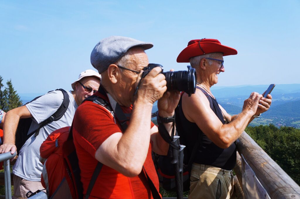 Trzech mężczyzn podziwia górskie widoki. Mężczyzna ubrany w czerwoną koszulkę i mężczyzna ubrany w czarną koszulkę i czerwony kapelusz robią zdjęcia. 
