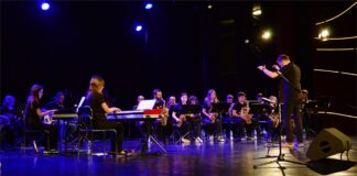 Obraz zawiera: Spojrzenie z boku sceny na kilkudziesięciu młodych uczestników warsztatów big bandowych siedzących przed pulpitami na scenie i grających na swoich instrumentach. Wspierają ich prowadzący warsztaty. Dyryguje Rafał Czarnacki.