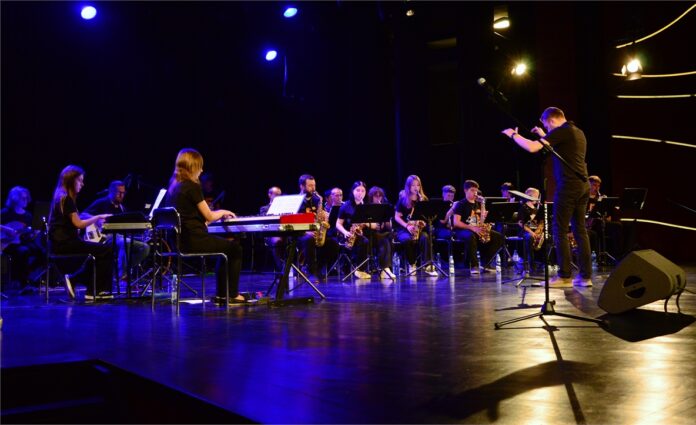 Finał V Letnich Warsztatów Big Bandowych_07 Obraz zawiera: Spojrzenie z boku sceny na kilkudziesięciu młodych uczestników warsztatów big bandowych siedzących przed pulpitami na scenie i grających na swoich instrumentach. Wspierają ich prowadzący warsztaty. Dyryguje Rafał Czarnacki.