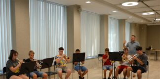 Obraz zawiera: Siedmioro młodych trębaczy siedzi na krzesłach i gra na trąbkach. Pomiędzy nimi stoi instruktor, który gestykulując prawą ręka tłumaczy im zasady gry.