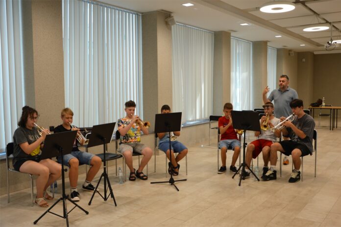 Warsztaty Big Bandowe_2 Obraz zawiera: Siedmioro młodych trębaczy siedzi na krzesłach i gra na trąbkach. Pomiędzy nimi stoi instruktor, który gestykulując prawą ręka tłumaczy im zasady gry.