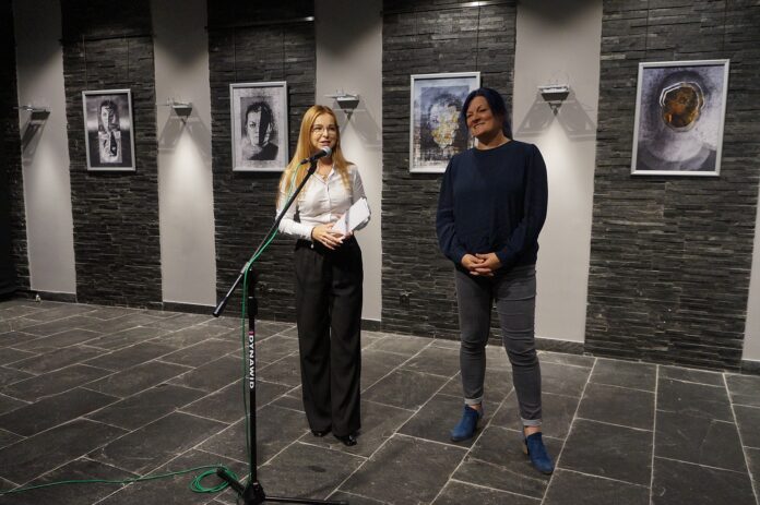 DSC05999 Dwie kobiety. Kobieta stojąca z lewej (Dyrektor Jasielskiego Domu Kultury), wita przybyłych gości. Ubrana jest w białą bluzkę i czarne spodnie. Kobieta z prawej strony (gość specjalny wernisażu), czeka na wystąpienie. Kobieta ubrana jest w granatową bluzkę i szare spodnie.