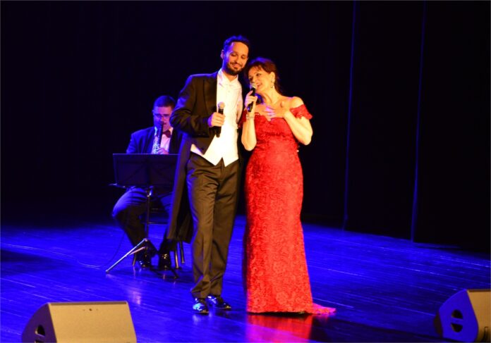 Koncert Jedz do Varazdin_5 Obraz zawiera: Tenor i sopranistka stoją w centralnej części sceny. Oboje trzymają w prawych dłoniach mikrofony. Mężczyzna ma na sobie ciemny frak, jasny surdut i koszulę. Kobieta szkarłatną suknię do ziemi. W tle z lewej strony za nimi klarnecista.