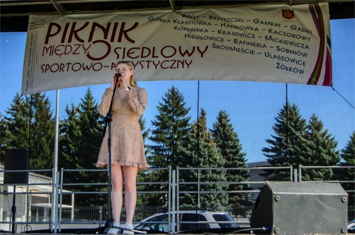 Piknik Międzyosiedlowy (323)_02 Obraz zawiera: Na scenie śpiewa do mikrofonu na statywie wokalistka JDK - Laura Gondek. Blondwłosa nastolatka trzyma oburącz mikrofon, ma lekko przymknięte oczy. Na sobie jasna sukienkę do kolan z rękawami.