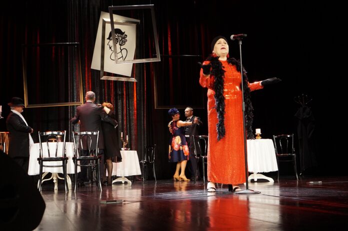 DSC06997 Na scenie stoi kobieta. Ubrana jest w długą, czerwoną suknię i czarny szal z piór. Głowę okrywa chusta w kolorze czarnym. Kobieta śpiewa do mikrofonu.