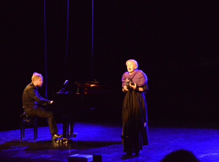 Znani jaślanie: Franciszka Platówna-Rotter, Adam Loret, Na scenie jasielskiego domu kultury występują starsza kobieta i młody mężczyzna. On ubrany na czarno akompaniuje na fortepianie, kobieta natomiast w czarnej sukni i fioletowym szalu, ozdobionym z przodu kwiatami wykonuje arie operowe. Kobieta ma jasnoblond włosy upięte w kok, mężczyzna modną, postrzępioną ciemnoblond czuprynę