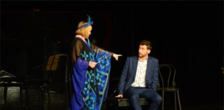 Obraz zawiera: Kobieta i mężczyzna na scenie. Kobieta w dziwnym nakryciu głowy stoi z lewej strony sceny zwracając się w prawą stronę. Lewą ręką wskazuje na siedzącego z prawej strony sceny na ławie do fortepianu mężczyznę, który patrzy na nią zaskoczony. W lewej ręce kobieta trzyma dyrygencką batutę.