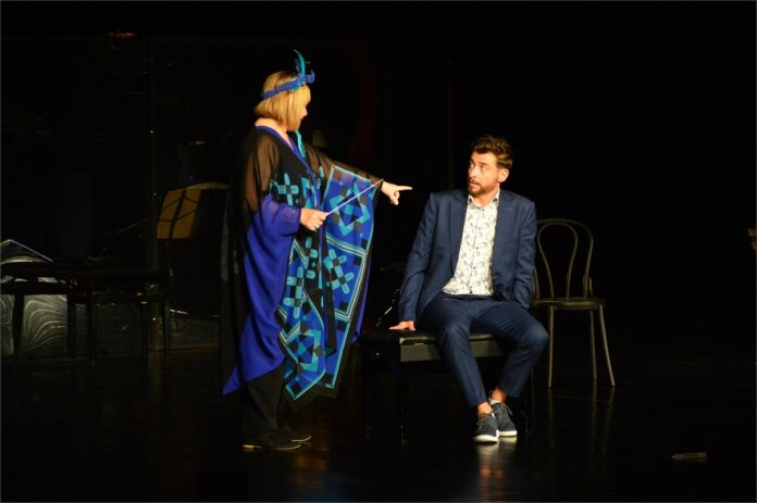 Obraz zawiera: Kobieta i mężczyzna na scenie. Kobieta w dziwnym nakryciu głowy stoi z lewej strony sceny zwracając się w prawą stronę. Lewą ręką wskazuje na siedzącego z prawej strony sceny na ławie do fortepianu mężczyznę, który patrzy na nią zaskoczony. W lewej ręce kobieta trzyma dyrygencką batutę.