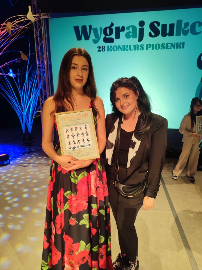 Na tle napisu Wygraj sukces stoi Karolina Polak w czarnej sukience w duże czerwone kwiaty, przed sobą trzyma dyplom. Obok niej niska kobieta, brunetka z długimi włosami, spiętymi w kucyk, ubrana na czarno w marynarkę i spodnie