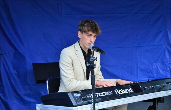 Obraz zawiera: Na plenerowej scenie Jasielskiego Domu Kultury, siedzi przed instrumentami klawiszowymi i gra na nich oraz śpiewa do stojącego przed nim na statywie mikrofonu - Mikołaj Majda, wokalista JDK, podopieczny trenerki wokalnej Ewy Grzebień.