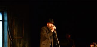 Obraz zawiera: Mężczyzna w kapeluszu, ciemnych spodniach, koszuli i kamizelce stoi na scenie JDK. Trzyma oburącz mikrofon na statywie śpiewa.