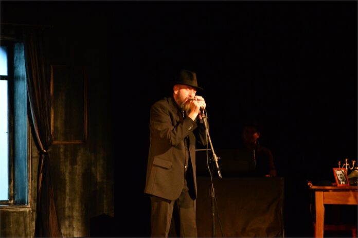 Cohen_3 Obraz zawiera: Mężczyzna w kapeluszu, ciemnych spodniach, koszuli i kamizelce stoi na scenie JDK. Trzyma oburącz mikrofon na statywie śpiewa.
