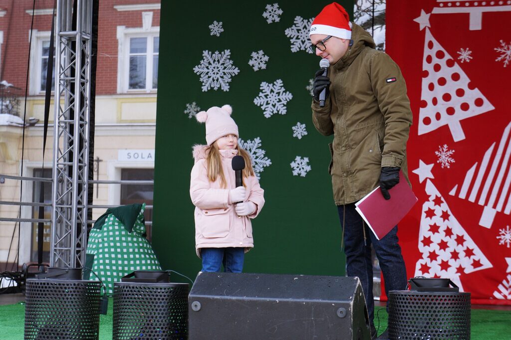 Na scenie stoją dwie osoby: mała dziewczynka i mężczyzna, Konrad Bilski, prowadzący tegoroczne wydarzenie. Konrad zapowiada występ dziewczynki.
