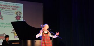 Obraz zawiera: Dziewczynka w kolorowej sukience w kratkę z rozłożonymi rękoma śpiewa do stojącego przed nią mikrofonu. W tle przy fortepianie instruktor śpiewu Ewa Grzebień, która akompaniuje młodej wokalistce.