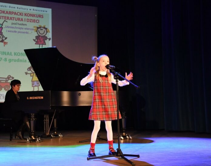 JaGODA RZESZÓW ikonka Obraz zawiera: Dziewczynka w kolorowej sukience w kratkę z rozłożonymi rękoma śpiewa do stojącego przed nią mikrofonu. W tle przy fortepianie instruktor śpiewu Ewa Grzebień, która akompaniuje młodej wokalistce.