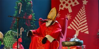 Obraz zawiera: Święty Mikołaj siedzi na tronie na scenie Jasielskiego Domu Kultury i przemawia do mikrofonu. Z jego lewej i prawej strony stoją paczki z prezentami.