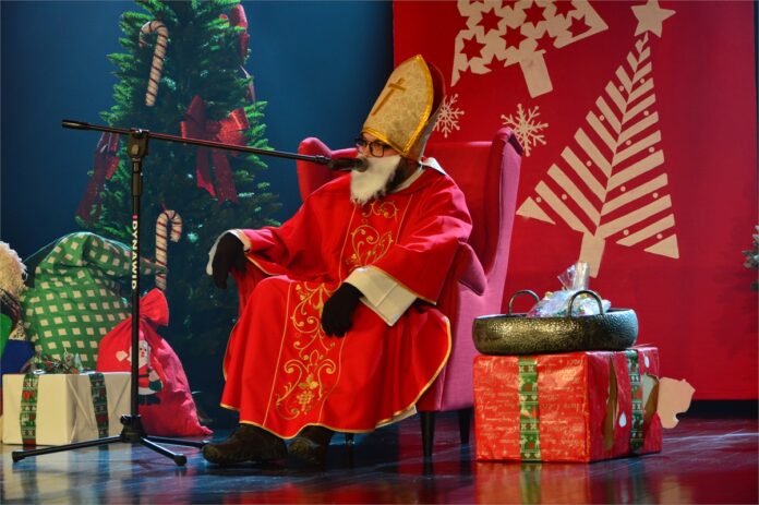 Mikolaj Caritas_4 Obraz zawiera: Święty Mikołaj siedzi na tronie na scenie Jasielskiego Domu Kultury i przemawia do mikrofonu. Z jego lewej i prawej strony stoją paczki z prezentami.