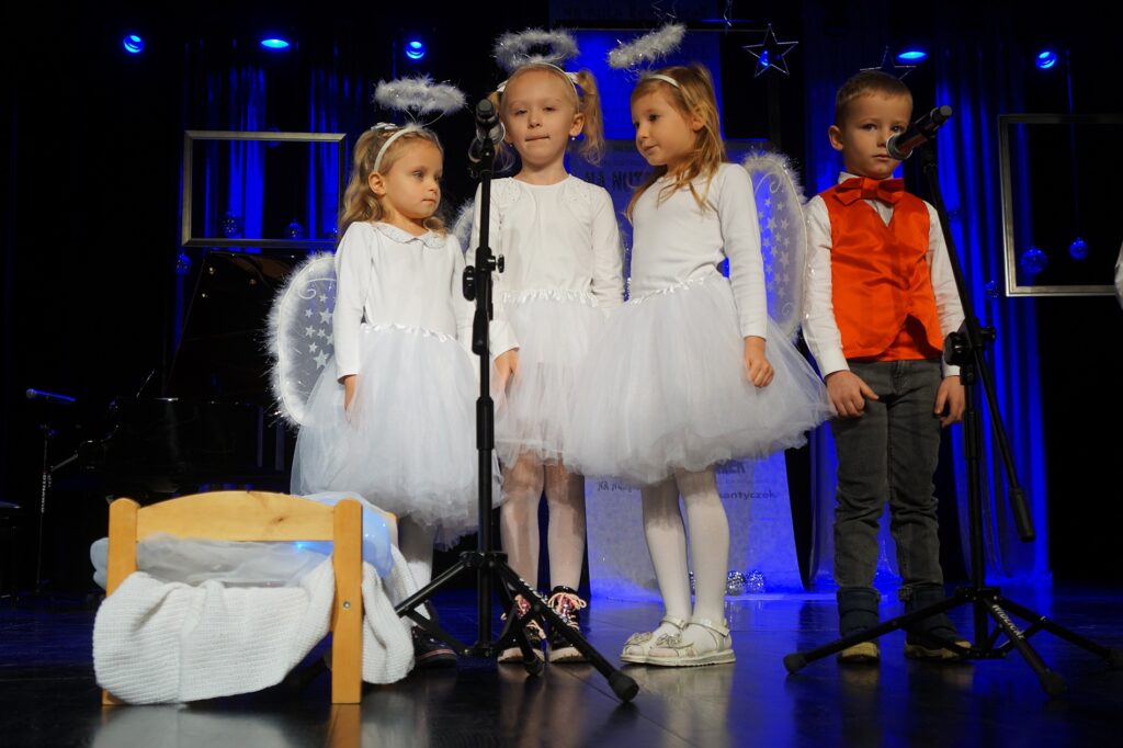 Czwórka dzieci występuje na scenie. Trzy dziewczynki występują w roli aniołków zaś chłopiec w roli śpiewaka.