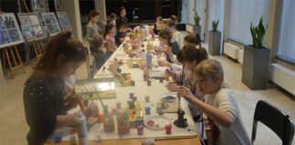 Świat glinianych dzieł Obraz zawiera: Dzieci siedzą po obu stronach długiego stołu i malują zrobione przez siebie gliniane figurki.