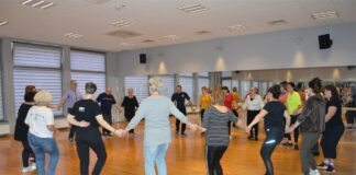 Tańce greckie w JDK Grupa osób w sali tanecznej JDK stojąc w kółku trzyma się za ręce i uczy się kroków tańca greckiego.